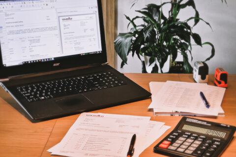 Biurko z laptopem, kalkulatorem i dokumentami przedstawia przygotowanie kosztorysu. W tle ładny, zielony kwiatek